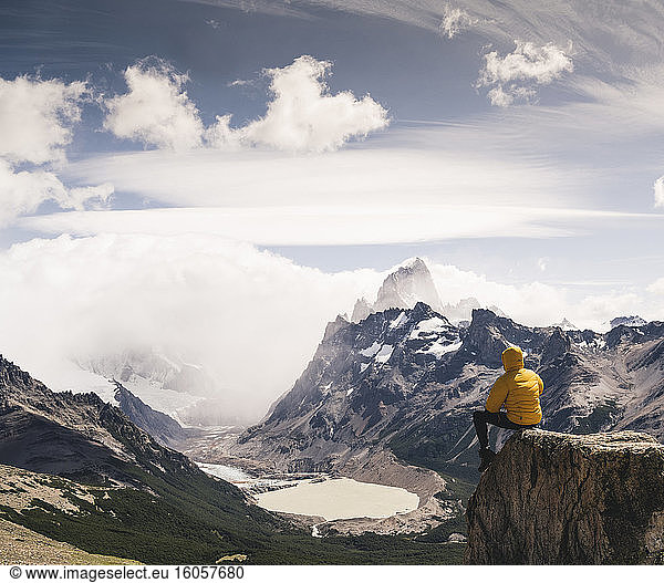 Mann schaut auf einen schneebedeckten Berg  während er auf einem Felsen gegen den Himmel sitzt  Patagonien  Argentinien
