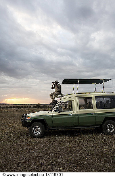 Mann schaut durch ein Fernglas  während er auf einem Geländewagen im Serengeti-Nationalpark sitzt