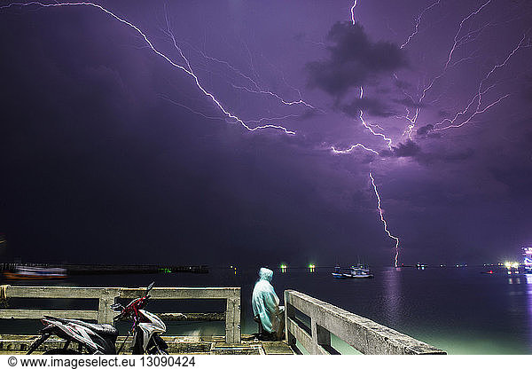 Mann sitzt am Pier am Meer gegen den Blitz am violetten Himmel