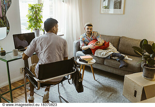 Mann sitzt auf einem Stuhl und spricht mit seinem Freund  der ein Smartphone im Wohnzimmer benutzt