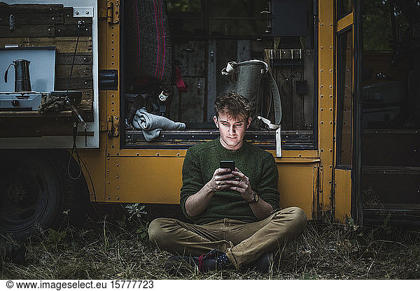 Mann sitzt im Schneidersitz  während er ein Smartphone gegen ein Wohnmobil beim Camping benutzt