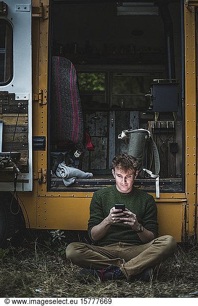 Mann sitzt in voller Länge im Schneidersitz  während er beim Camping mit seinem Smartphone gegen ein Wohnmobil fährt