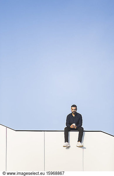 Mann telefoniert  während er auf einem Bauwerk vor blauem Himmel sitzt