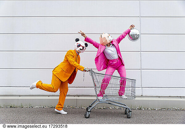 Mann und Frau in farbenfrohen Anzügen und Tiermasken  die mit Einkaufswagen und Discokugel herumspielen