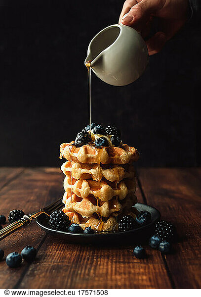 Maple syrup being poured over stack of belgian waffles with berries.