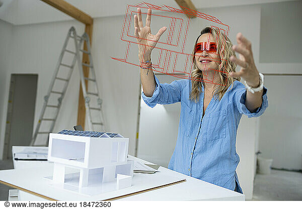 Mature architect wearing virtual reality simulator gesturing and examining house model at site
