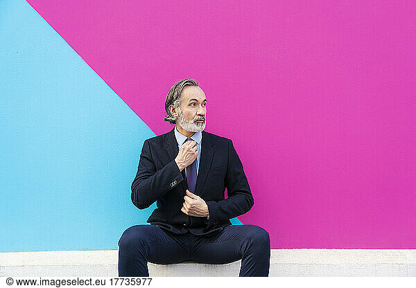 Mature businessman adjusting tie sitting in front of pink and blue wall