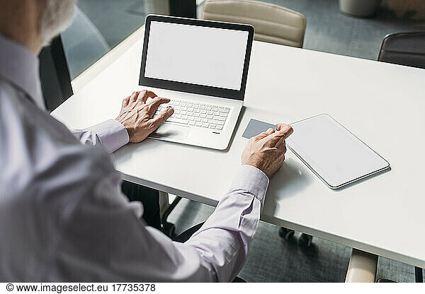 Mature businessman holding credit card making online payment through laptop in office