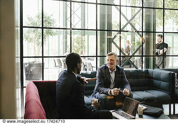Mature businessman looking at male professional discussing while sitting on sofa at convention center
