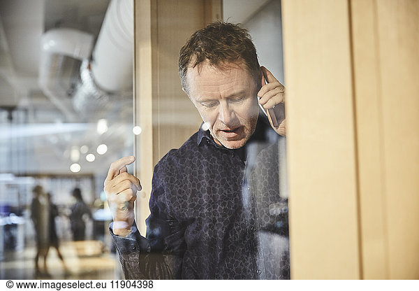 Mature businessman talking on mobile phone seen from glass at office