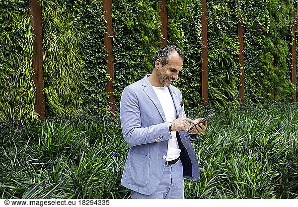 Mature businessman text messaging on mobile phone standing in front of hedge