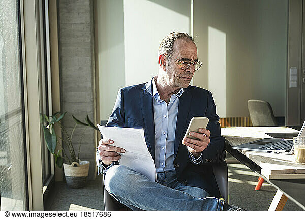 Mature businessman using smart phone sitting with document in office