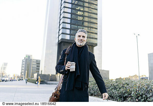 Mature businessman walking with coffee cup at footpath