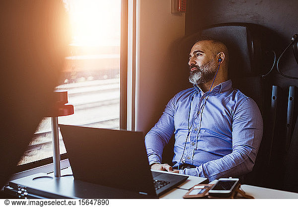 Mature businessman wearing headphones looking away while using laptop in train