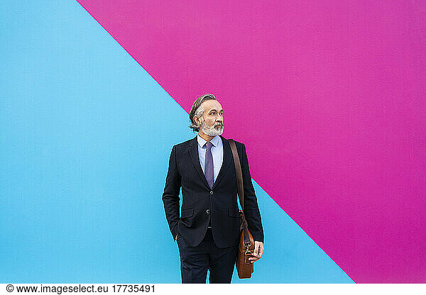 Mature businessman with bag standing in front of pink and blue wall