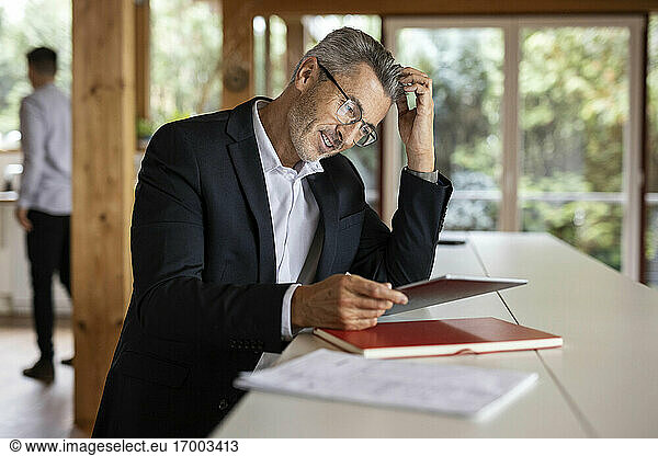 Mature businessman with hand in hair using digital tablet while sitting by table at home