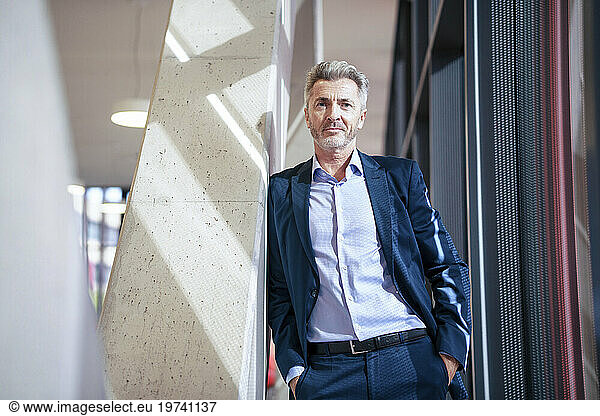 Mature businessman with hands in pockets standing near column