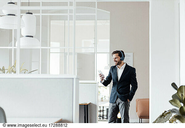 Mature businessman with headphones using mobile phone while standing in office