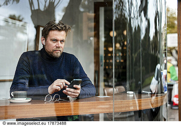 Mature businessman with mobile phone sitting in coffee shop seen through glass window