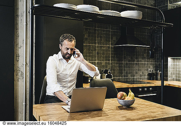 Mature businessman working in kitchen  using laptop  talking on the phone