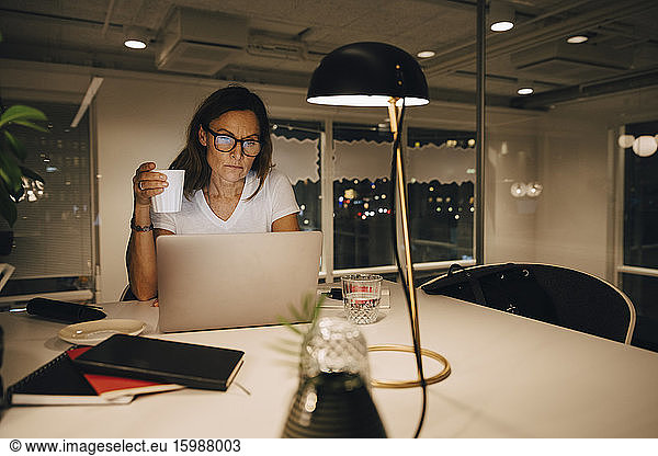Mature businesswoman holding coffee cup while working late at illuminated creative workplace
