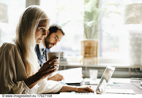 Mature businesswoman holding coffee mug using laptop by businessman at desk in office