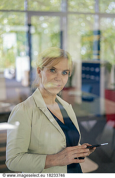 Mature businesswoman holding mobile phone at office seen through glass