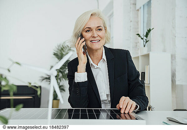 Mature businesswoman sitting with solar panel and talking on mobile phone at desk