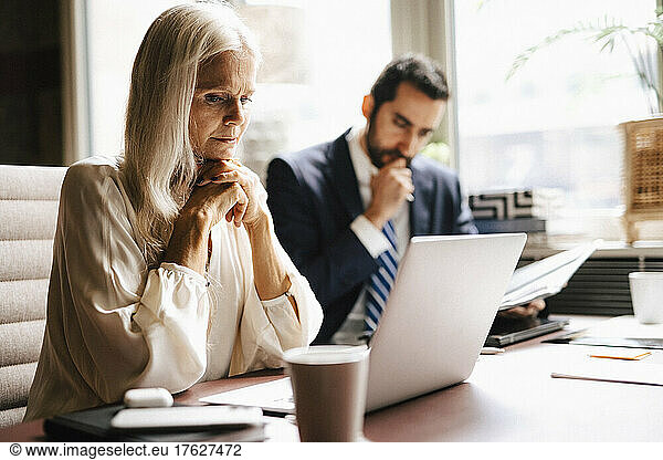 Mature businesswoman staring at laptop by businessman at desk in office