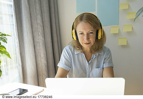 Mature businesswoman wearing wireless headphones using laptop working in home office