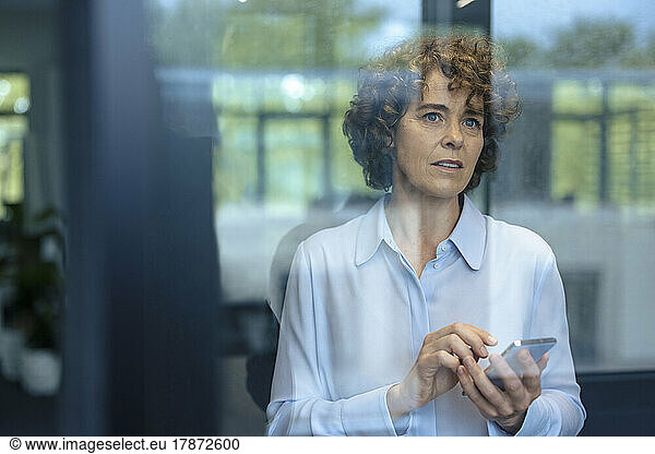 Mature businesswoman with curly hair holding smart phone seen through glass