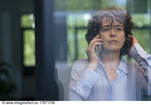 Mature businesswoman with hand in hair talking on smart phone seen through glass