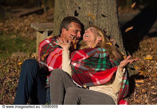 Mature couple sitting against tree trunk throwing autumn leaves at night