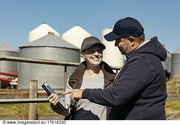 Mature couple smiling at each other standing by a fence with grain bins in the background  checking a tablet computer to manage their farm; Alcomdale  Alberta  Canada