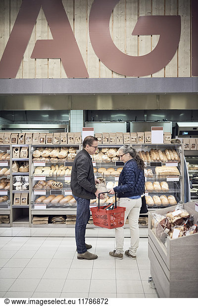 Mature couple standing with basket against bread rack at supermarket