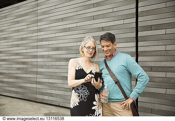 Mature couple using smart phone while standing against wall