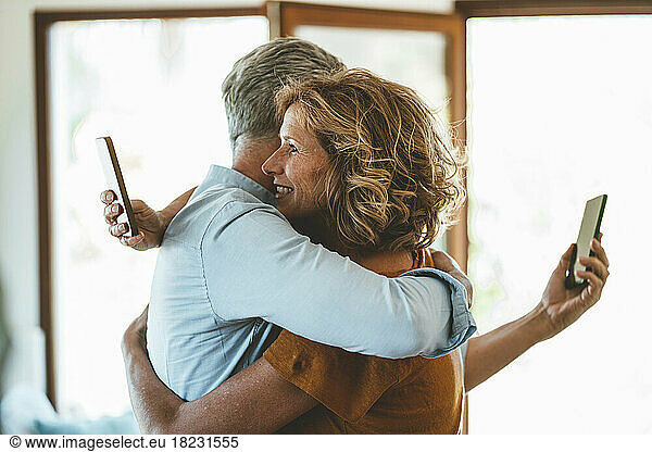 Mature couple with smart phones embracing each other at home
