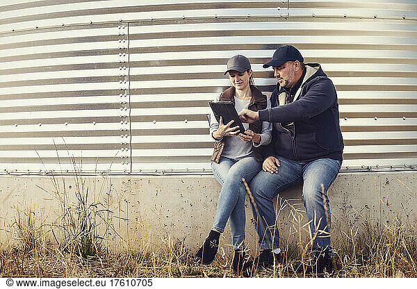 Mature couple working on their farm  sitting at on edge of a grain bin while consulting their tablet computer; Alcomdale  Alberta  Canada