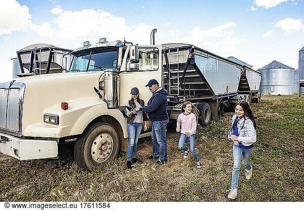 Mature couple working on their farm  standing next to a diesel transport truck and consulting their tablet computer while their two daughters have fun running around next to them; Alcomdale  Alberta  Canada