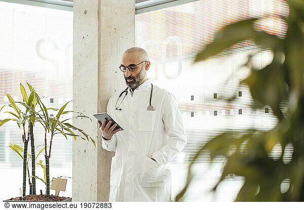 Mature doctor using tablet PC and leaning on column