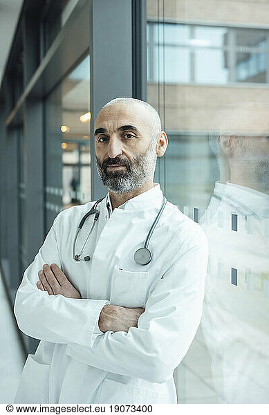Mature doctor with arms crossed leaning on glass window