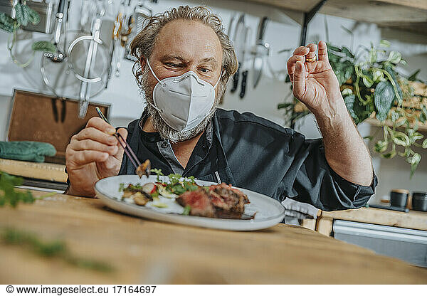 Mature expertise wearing face mask gesturing OK sign while garnishing dish in kitchen