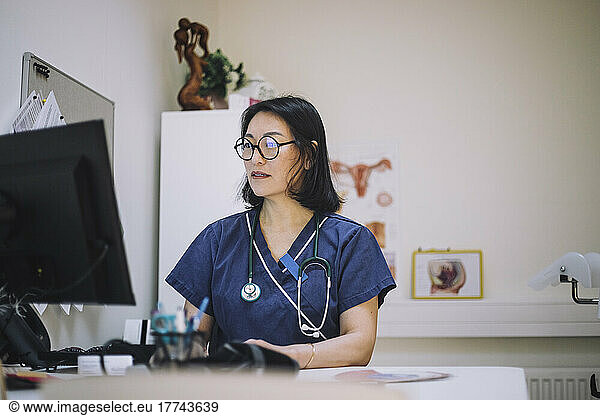 Mature female doctor wearing eyeglasses using computer sitting at desk in hospital