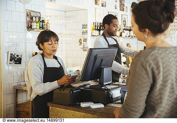 Mature female employee billing customer standing at checkout counter in delicatessen