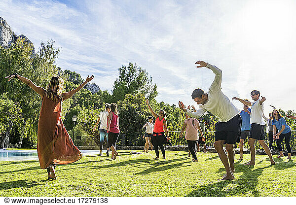Mature female fitness instructor dancing with tourists at health retreat on grass against sky during sunny day