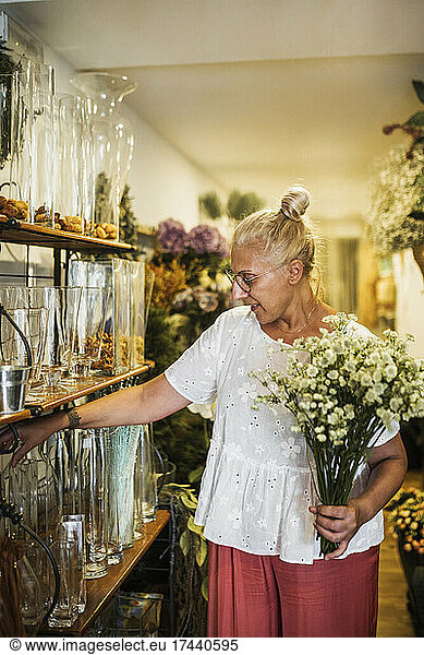 Mature female florist with flowering plant choosing vase in flower shop