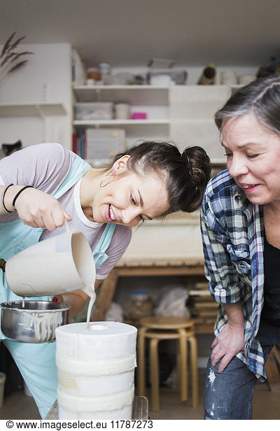 Mature female potter bending while looking at smiling young employee pouring clay from pitcher in vase at workshop