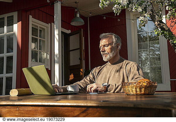 Mature freelancer working on laptop sitting outside house
