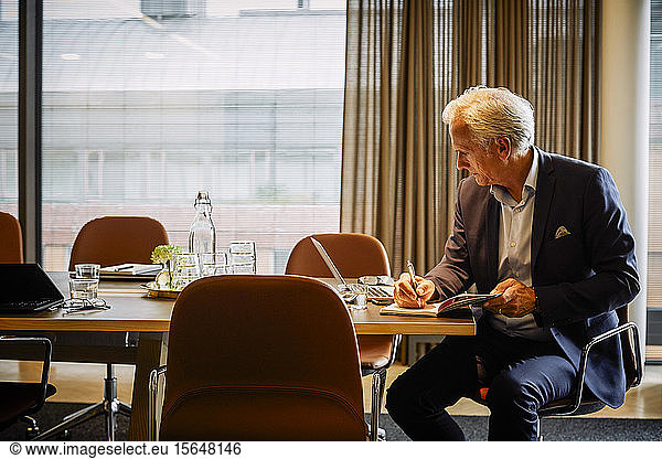 Mature legal professional writing in diary while using laptop at conference table in board room