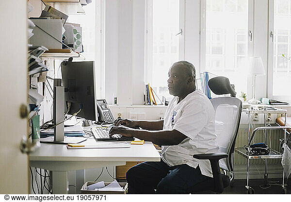 Mature male doctor working on computer while sitting at desk in clinic